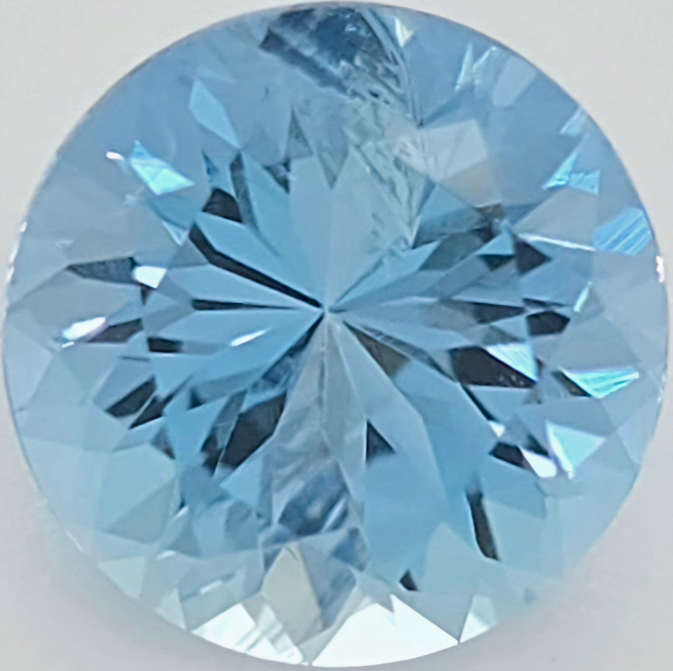 A round brilliant-cut light blue gemstone, showing detailed faceted reflections on a plain light background.
