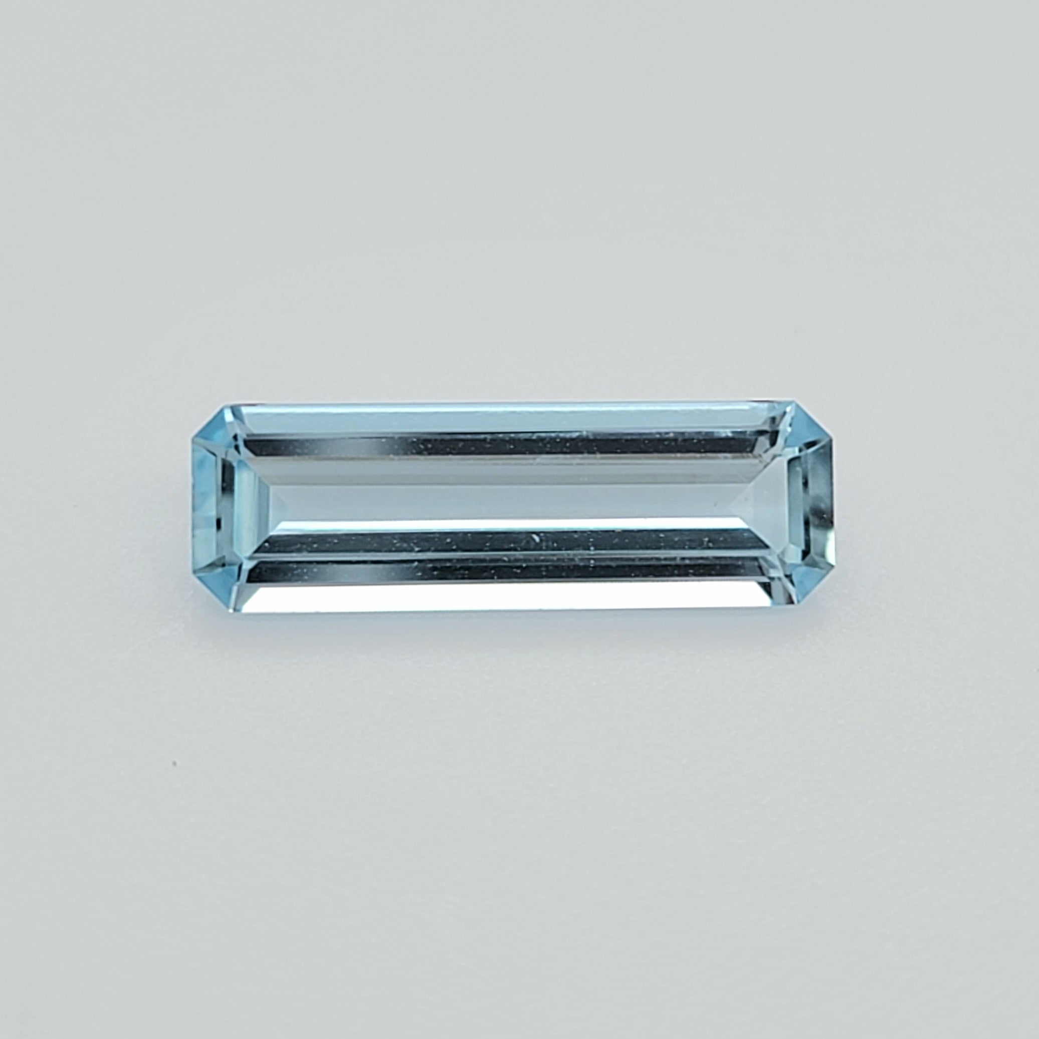 A rectangular emerald-cut light blue gemstone with a faceted, reflective surface, centered on a plain light gray background.