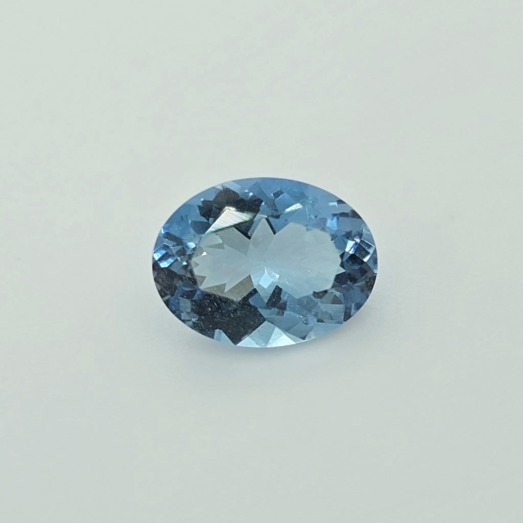 An oval-cut light blue gemstone with faceted cuts, centered on a plain light gray background.