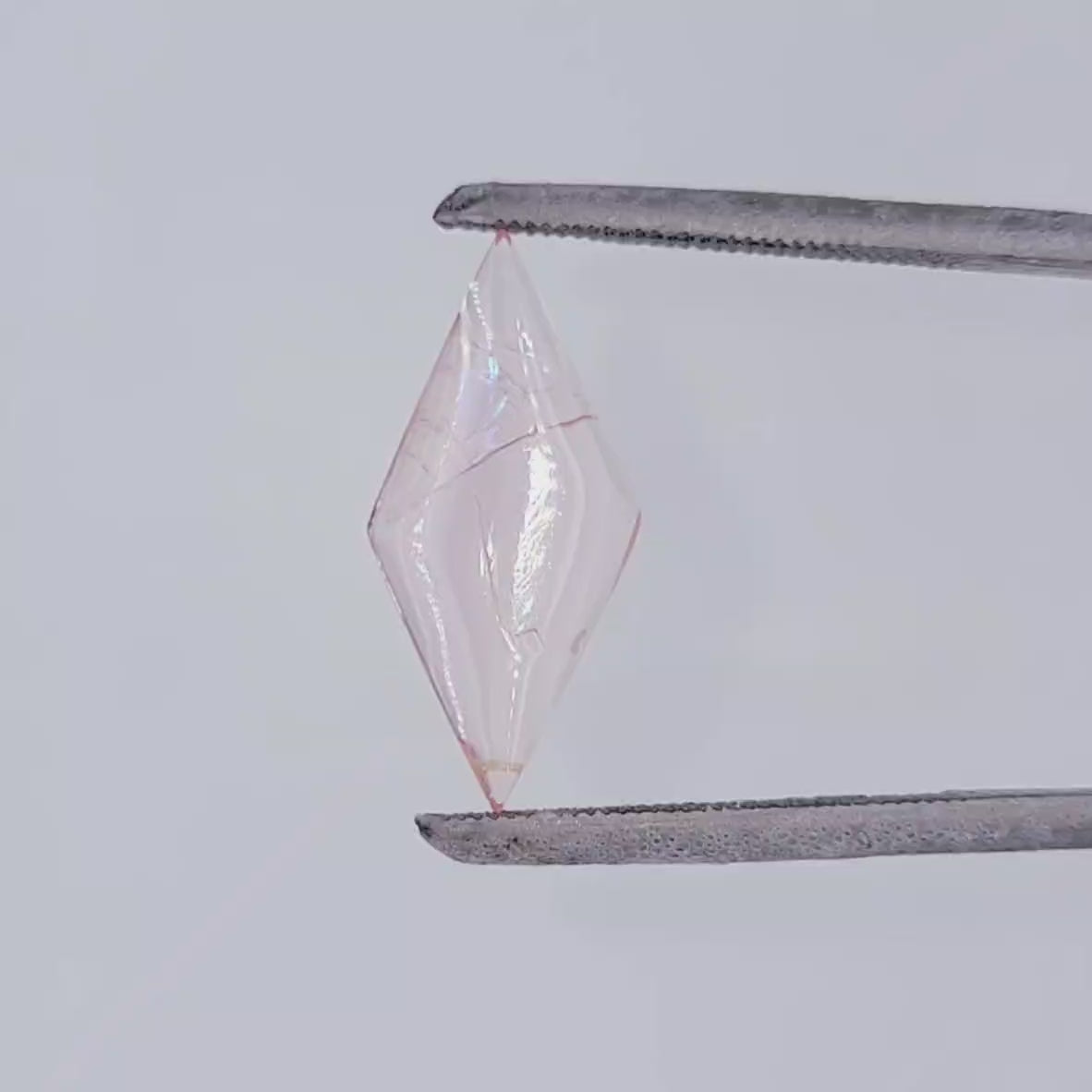 Video of a light pink diamond shaped gemstone with natural inclusions in tweezers against a white background