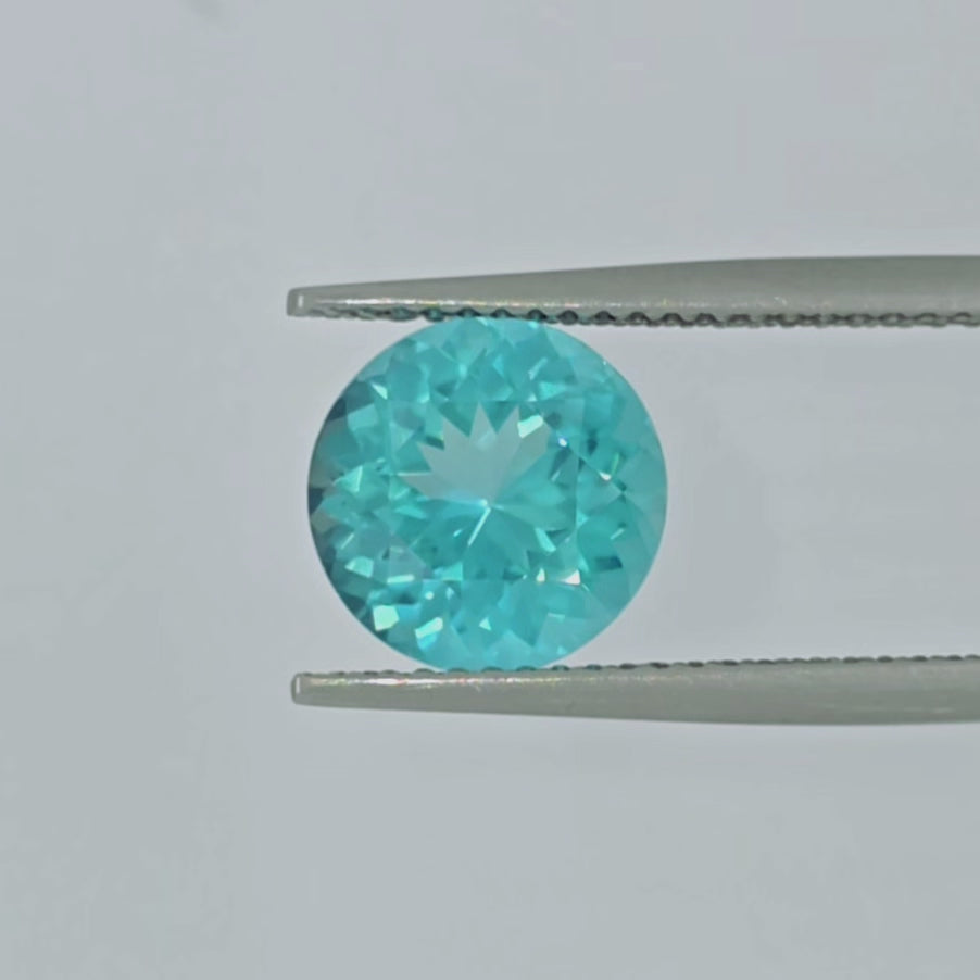 A video of a round greenish blue gemstone in tweezers against a pale gray background.