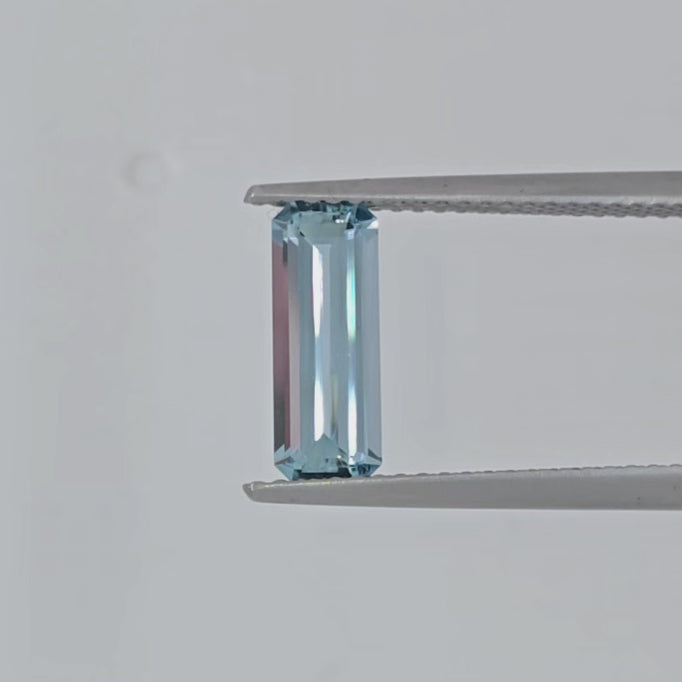 Video of a rectangular emerald cut light blue gemstone in tweezers against a white background