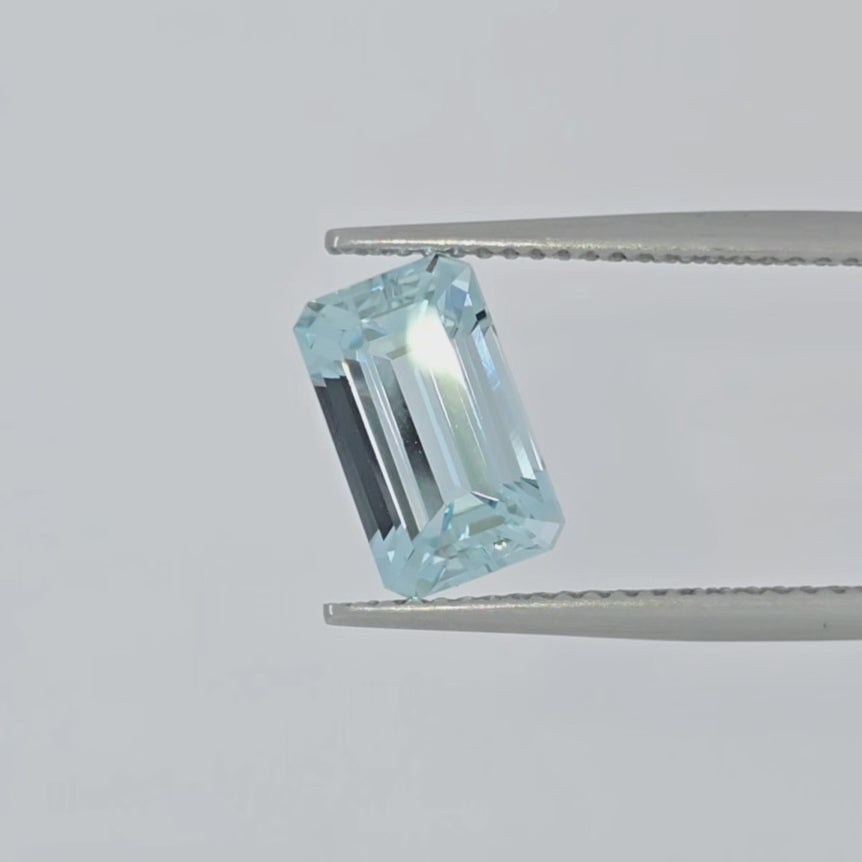 Video of a faceted emerald cut light blue gemstone in tweezers against a white background