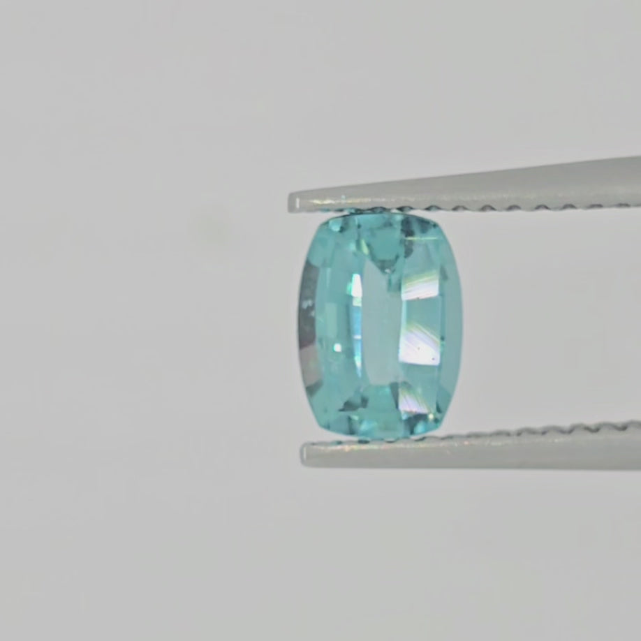 Video of A single cushion-cut light blue gemstone with a faceted, polished finish, shown in tweezers on a plain light gray background.