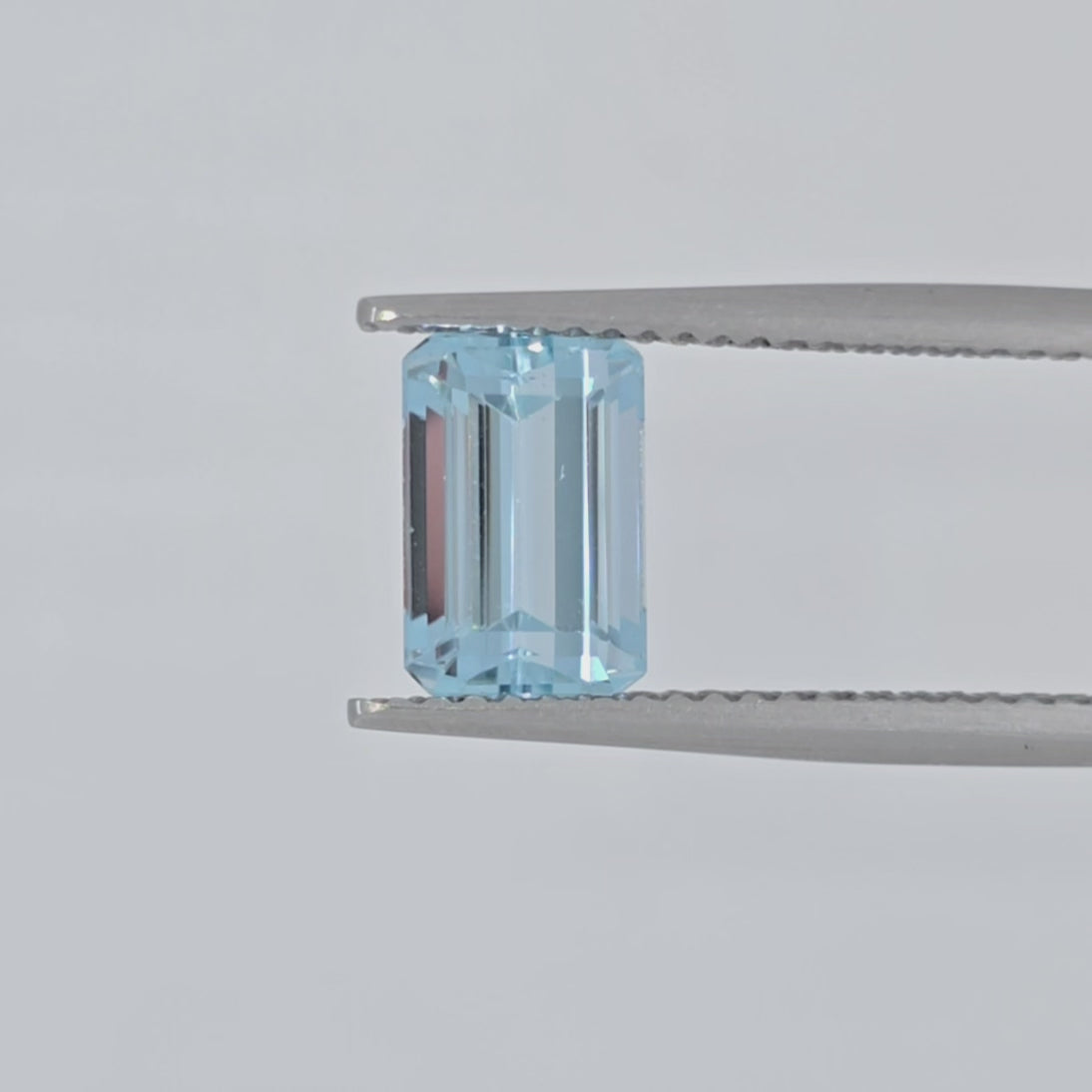 Video of a light blue emerald cut faceted gemstone in tweezers against a plain white background