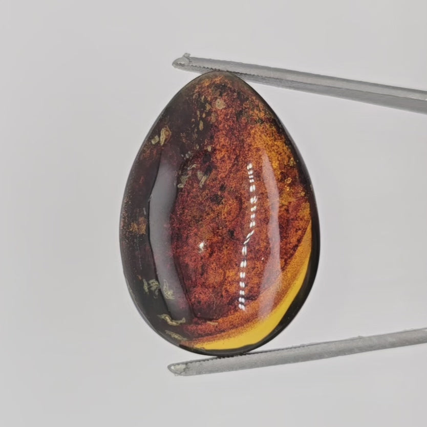 Video of teardrop shaped brownish cabochon with numerous inclusions in tweezers against a light background.