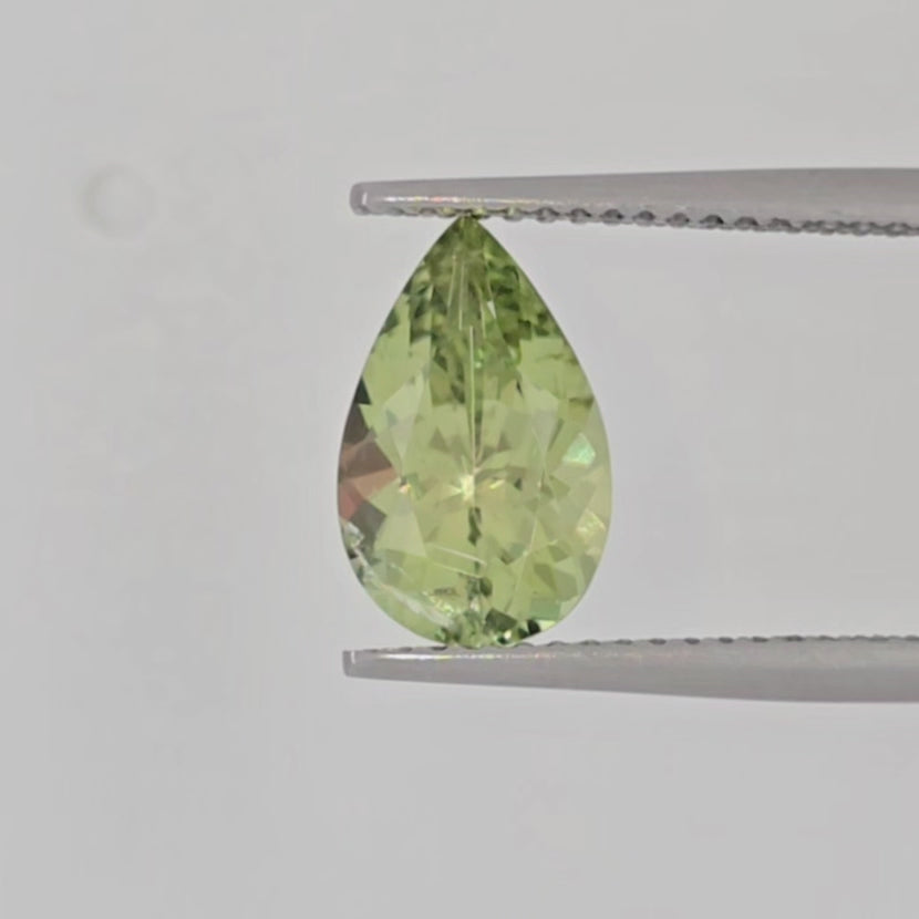 A video of a faceted pear-cut gemstone with a lighter lime green color, displayed in tweezers against a clean, off-white background.