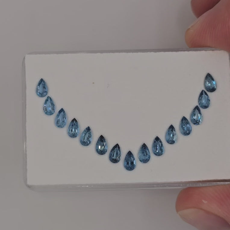 Video of 15 blue pear shaped gemstones in a curved line on a white rectangle pad held in fingers against a white background