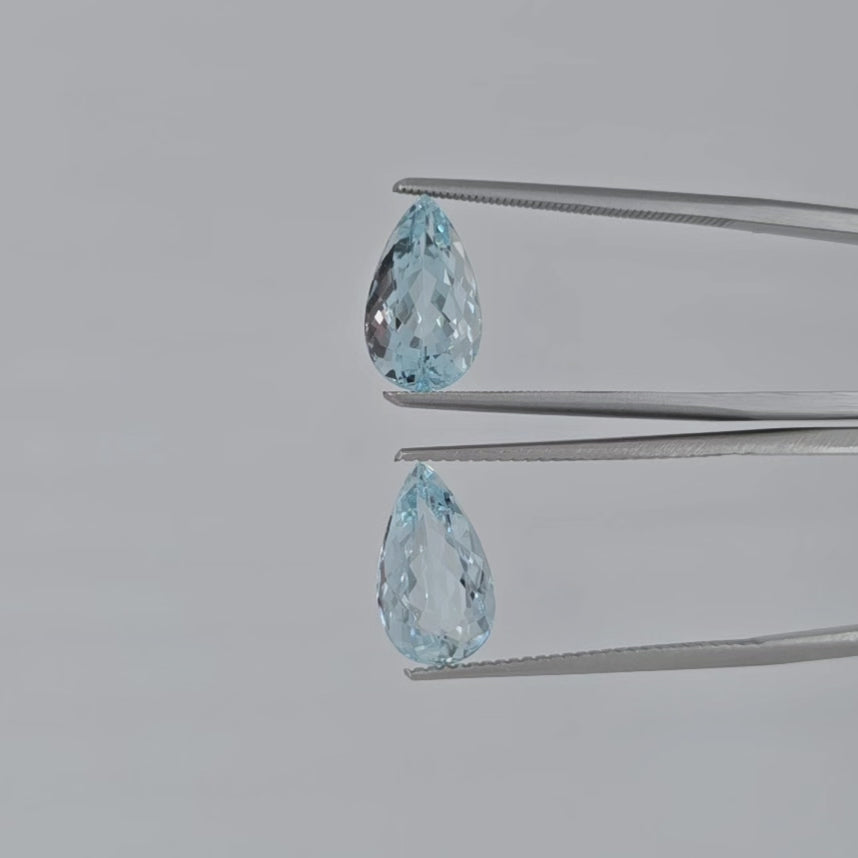 Video of two pear shaped faceted light blue gemstones in tweezers against a light gray background