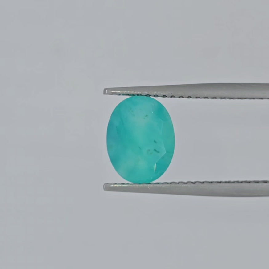 Video of a faceted oval greenish blue gemstone with natural inclusions, shown on a plain white background displayed in tweezers