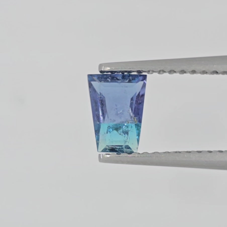 Video of trapezoid shaped gemstone with a blue to turquoise gradient, displayed on a neutral light gray background shown in tweezers