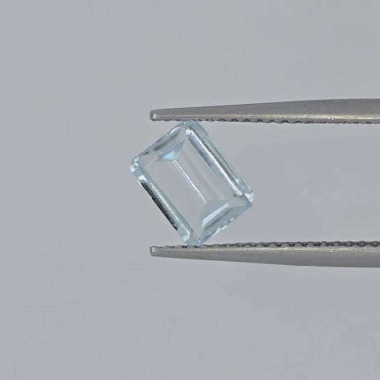 Video of a pale blue emerald cut faceted gemstone in tweezers against a pale gray background