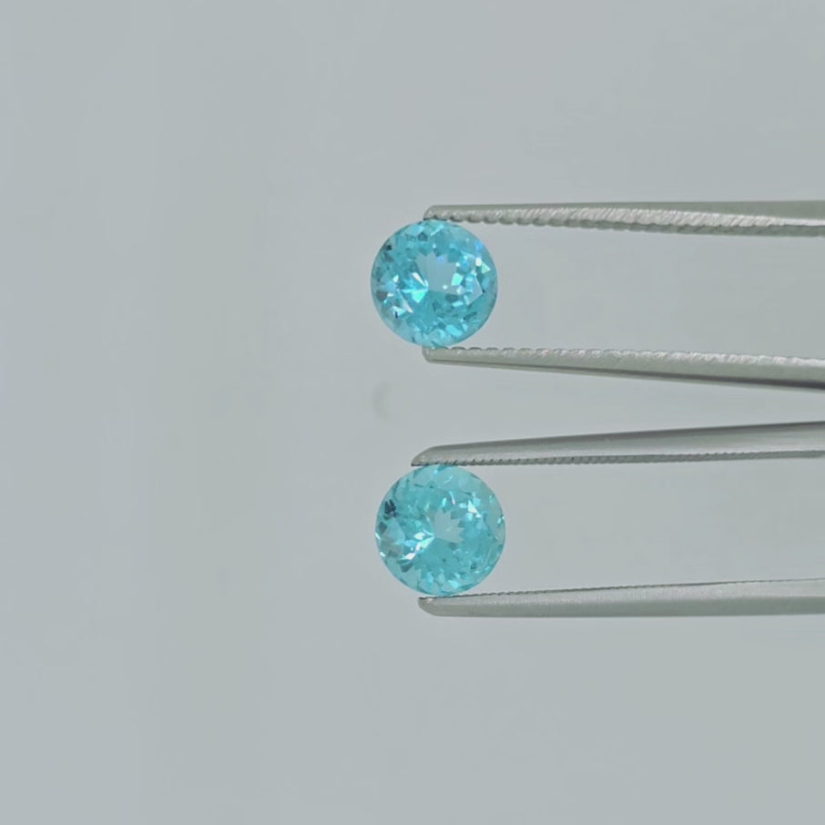 video of Two round brilliant-cut light blue gemstones displayed side-by-side on a clean, light gray background being shown in tweezers