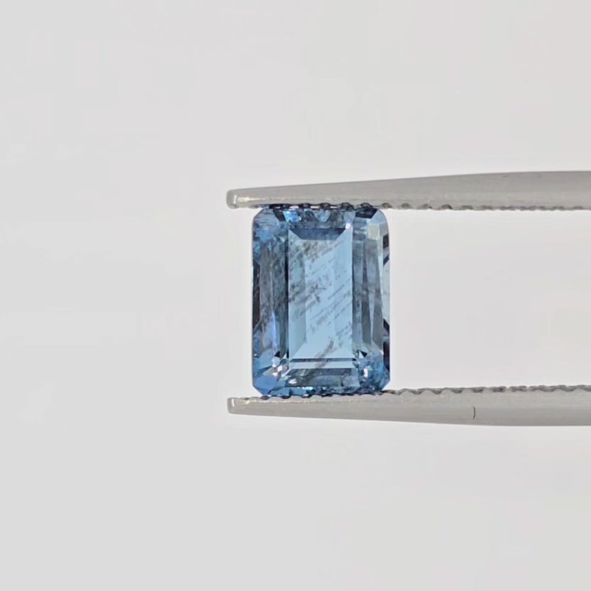 Video of an emerald cut faceted blue gemstone in tweezers against a plain white background