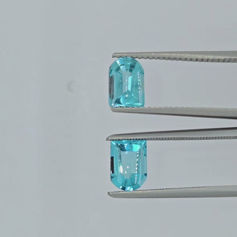 A video of two rounded rectangular cut blue gemstones displayed in tweezers side by side on a plain white surface.