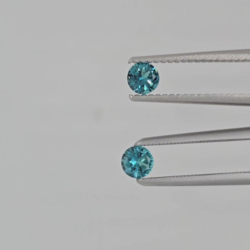 Video of two round brilliant cut greenish blue gemstones in tweezers against a light gray background