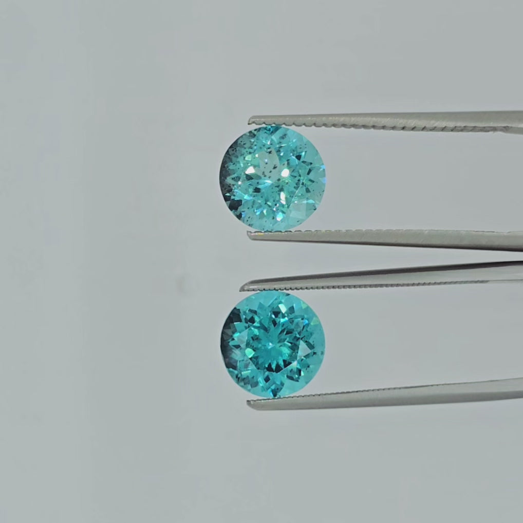 Video of a pair of round blue gemstones in tweezers being shown against a light gray background.