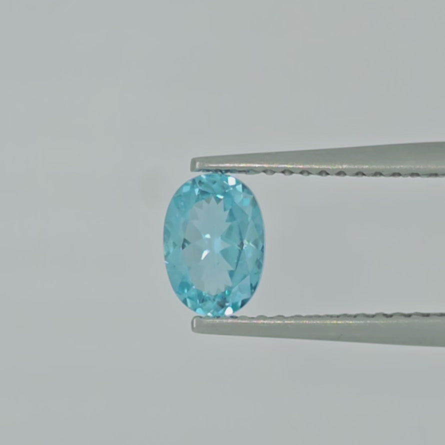 Video of a faceted oval greenish blue gemstone centered on a plain light gray background shown in tweezers