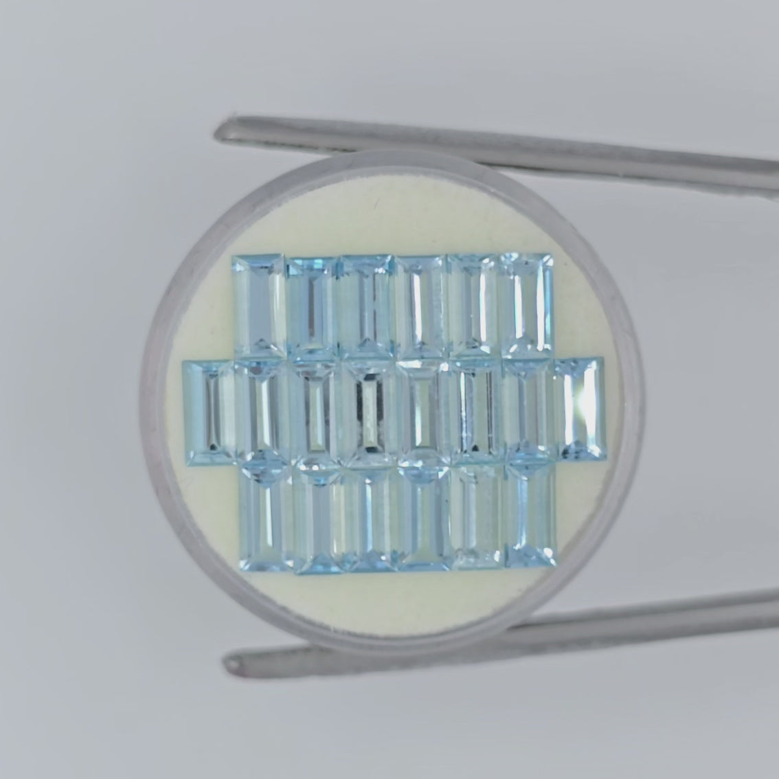 video of twenty rectangle cut light blue faceted stones on an off-white circular pad in tweezers on a white background