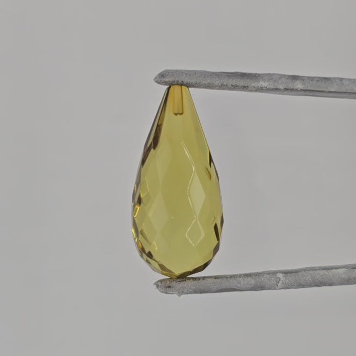 Video of a teardrop shaped faceted greenish yellow briolette in tweezers against a light gray background.