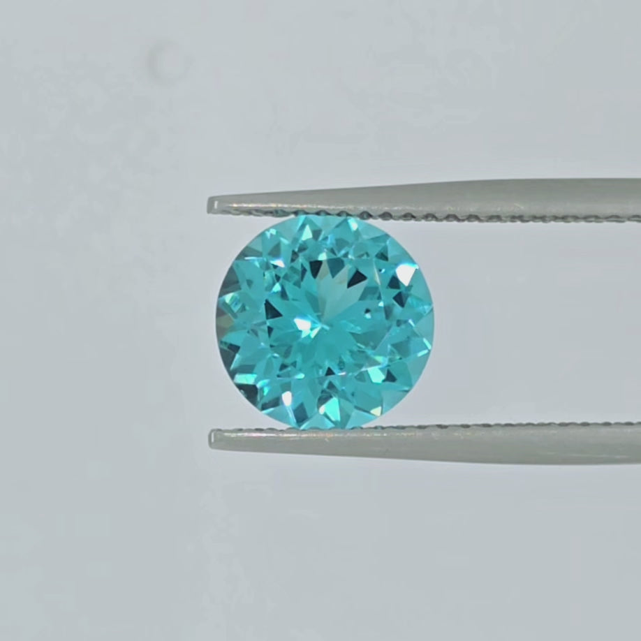 Video of a round greenish blue round gemstone in tweezers on a clean pale gray background.