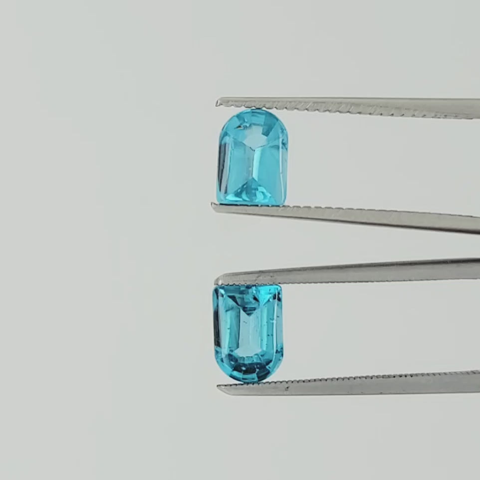 A video of a pair of light blue faceted gemstone cabochons with a rounded top and rectangular body, displayed in tweezers side-by-side on a plain light background.