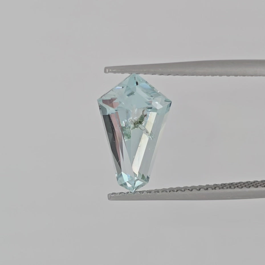 Video of a kite shape light blue gemstone with green inclusion in tweezers on a white background