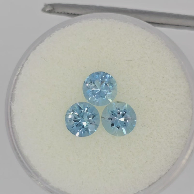 Video of three round faceted light blue gemstones on an off-white round pad in tweezers against a white background