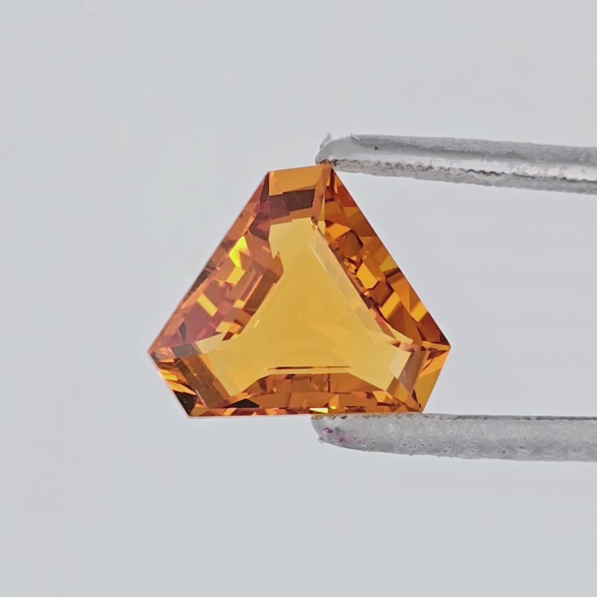 Video of a triangular cut orange gemstone in tweezers against a plain light gray background.