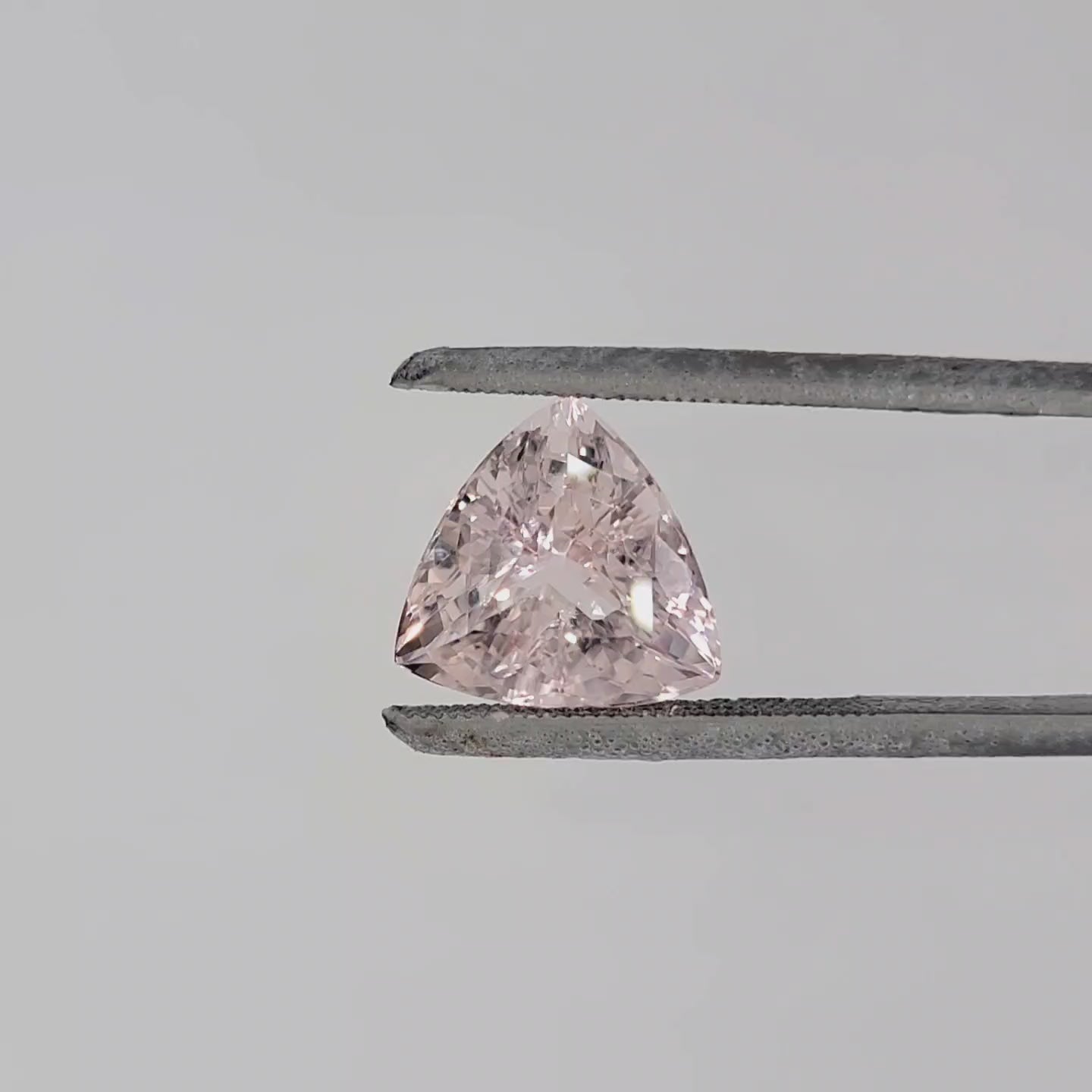 Video of a triangular shaped faceted pink gemstone in tweezers against a pale gray background